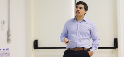 Um homem moreno, com bigode e cavanhaque escuros bem definidos, está em pé, olhando para a direita e levemente para cima. Ele veste uma camisa social de mangas compridas com listras finas azuis e brancas, calças escuras e um cinto marrom com fivela prateada. Sua mão direita está no bolso e a esquerda segura um objeto preto pequeno, possivelmente um controle remoto ou um apontador laser. Ao fundo, há uma parede branca com uma barra horizontal preta e parte de uma porta à direita. À esquerda, parcialmente visíveis, estão algumas garrafas de água sobre uma superfície branca.Webinar sobre investimento anjo para startups.