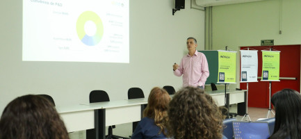A imagem mostra um homem em pé, de frente para uma plateia, em uma sala que parece ser um auditório ou sala de aula. Ele está gesticulando e parece estar apresentando algo, possivelmente explicando os gráficos e textos projetados na tela ao fundo. A plateia é composta por diversas pessoas sentadas em cadeiras azuis, voltadas para o apresentador e a tela. Algumas pessoas na plateia parecem estar olhando para a tela ou para o apresentador, enquanto uma pessoa na frente à direita parece estar olhando para um laptop. Fim da descrição.